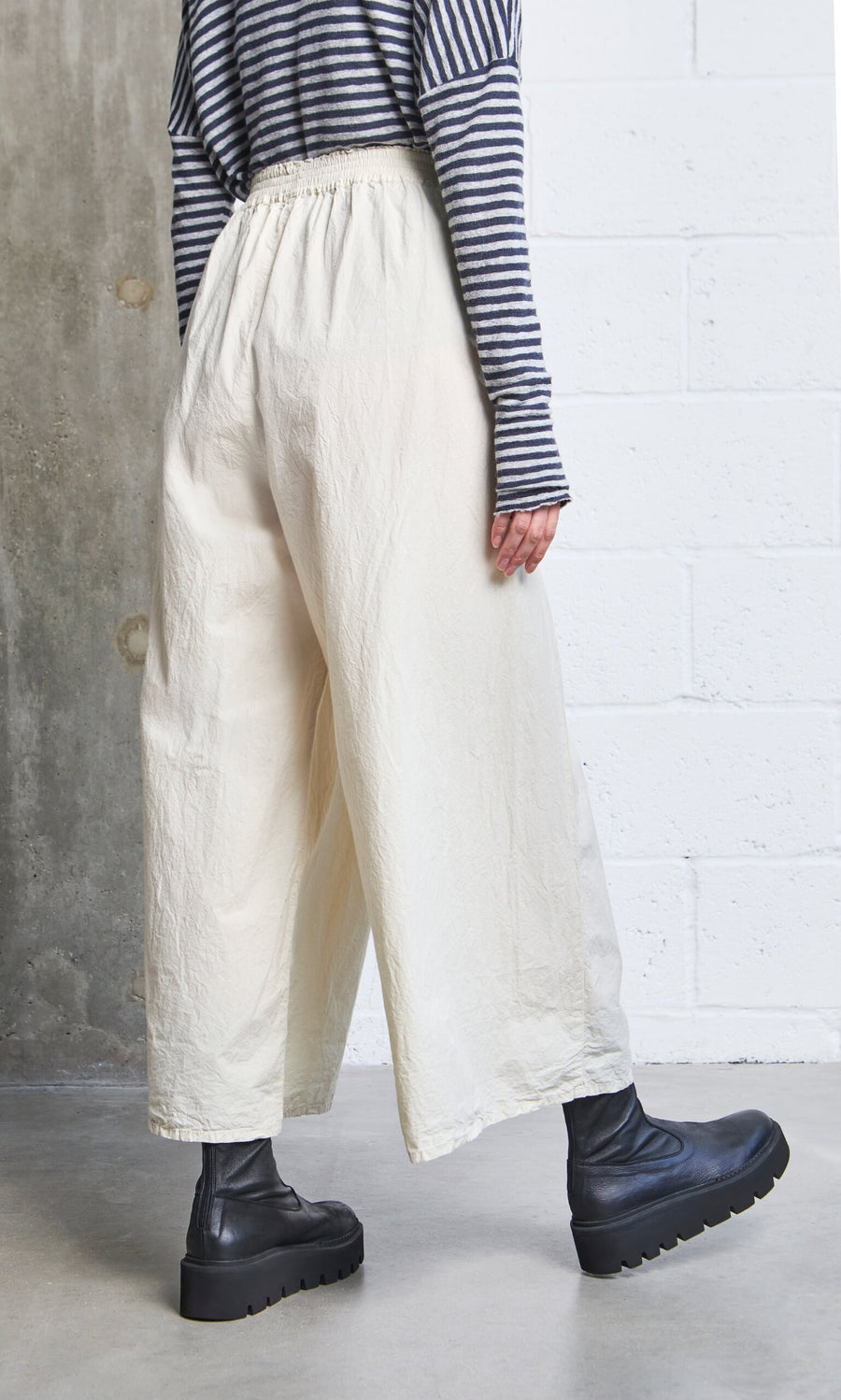 Plümo- A person in Jung Pants, a striped long-sleeve top, and black platform boots stands by a white brick wall.