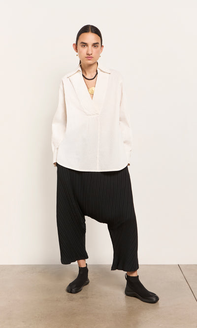 Plümo  Person in a loose Kaburi Shirt, black drop-crotch pants, black socks, and necklace stands against a plain wall.