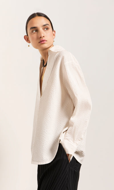 Plümo  Person in the Kaburi Shirt with cutaway collar, black pants, looking sideways against a light background.