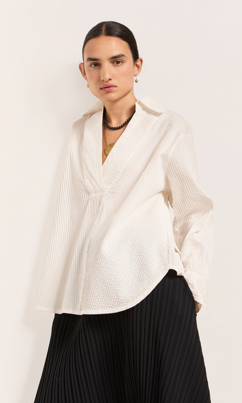 Plümo  Person wears a Kaburi Shirt, a textured white blouse with cutaway collar, and a black pleated skirt against plain background.