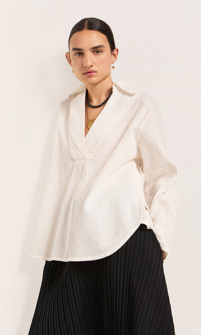 Plümo  Person wears a Kaburi Shirt, a textured white blouse with cutaway collar, and a black pleated skirt against plain background.
