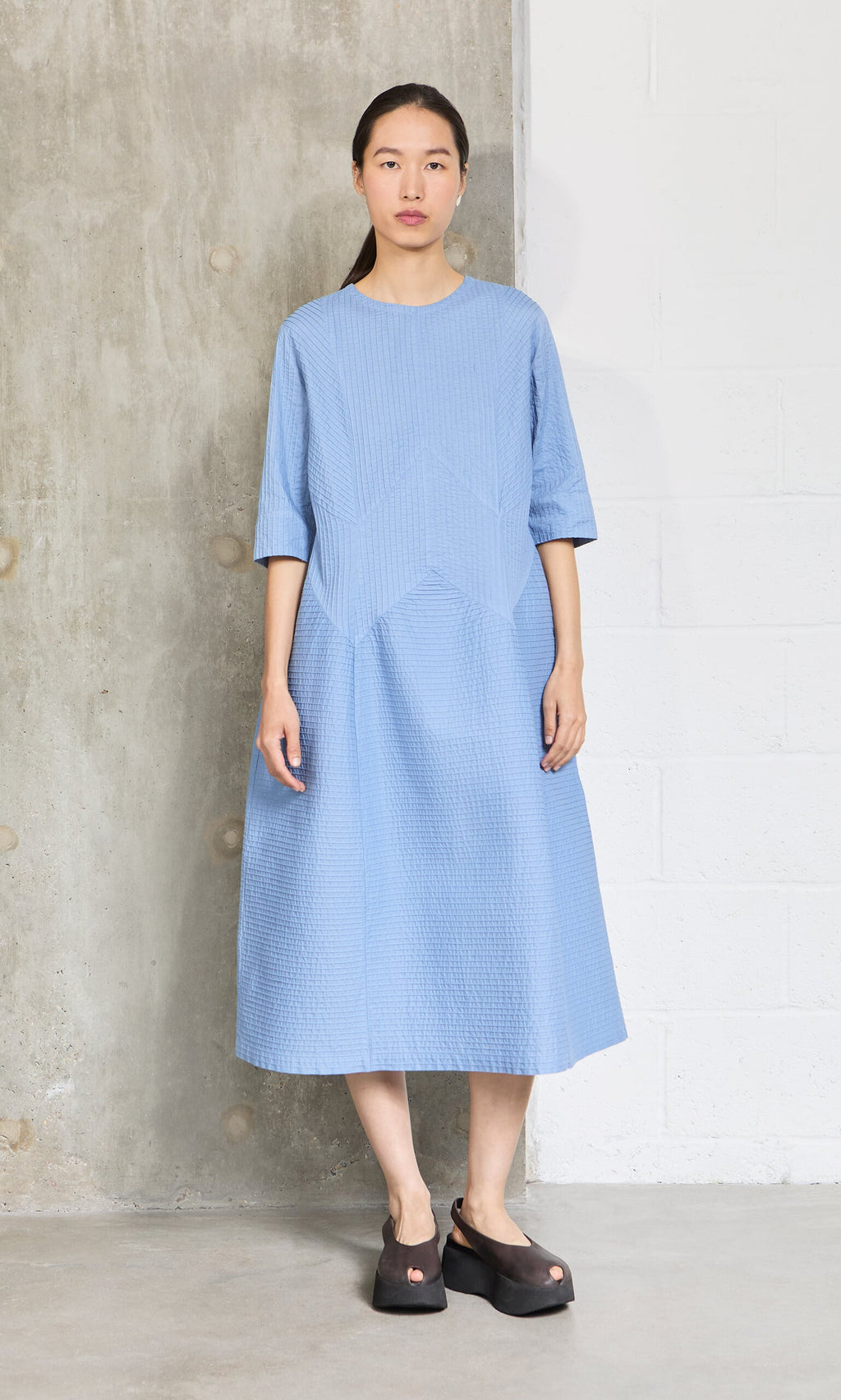 Plümo- Woman in the Kai Pleated Dress sky blue cotton with pintuck panels and black platform shoes stands by a concrete wall.