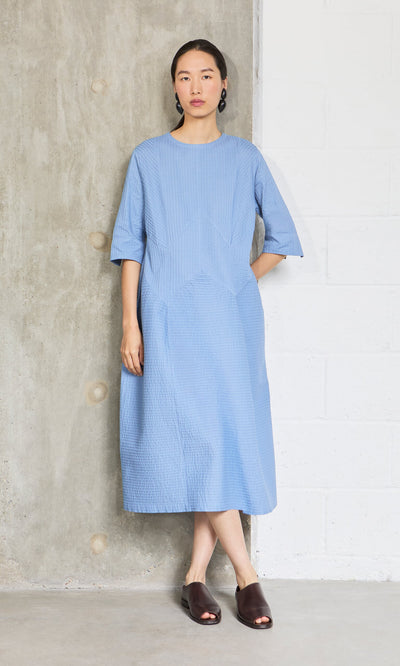 Plümo- Woman in a sky blue Kai Pleated Dress by Yacco Maricard stands against a concrete and brick wall.