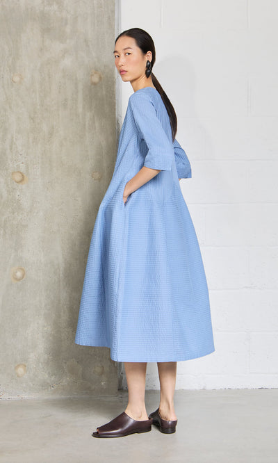 Plümo- Woman in a sky blue Kai Pleated Dress stands sideways against a concrete and white brick wall.