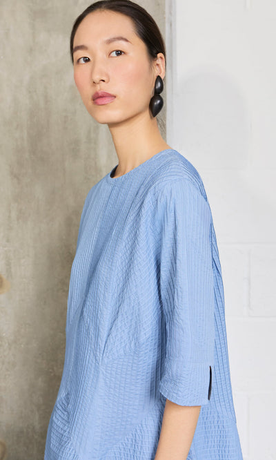 Plümo- Woman with slicked-back hair in a sky blue Kai Pleated Dress, standing by a concrete wall.