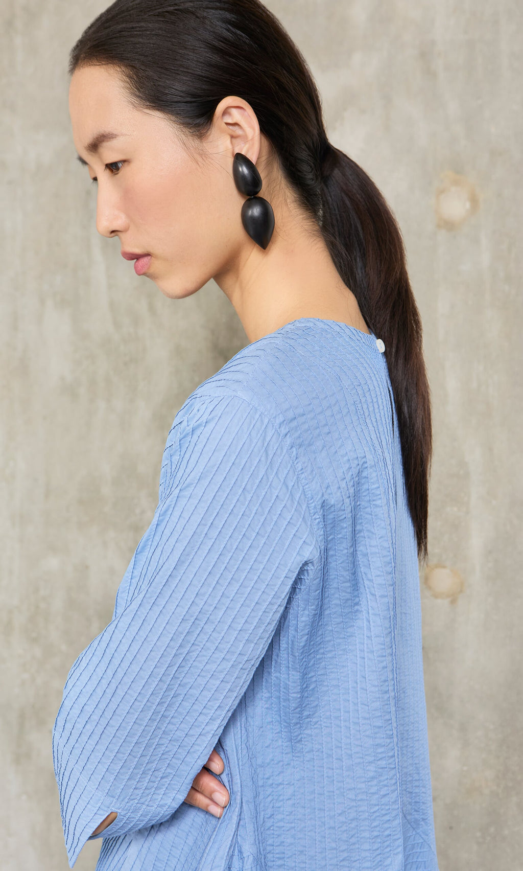 Plümo- Woman in profile with long dark ponytail, wearing the Kai Pleated Dress in sky blue cotton by Yacco Maricard.