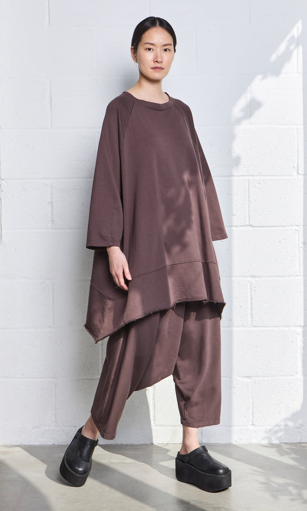 Plümo- Wearing Sybel Pants, a person stands against a white brick wall in soft lighting and black platform shoes.
