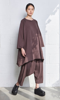Plümo- Wearing Sybel Pants, a person stands against a white brick wall in soft lighting and black platform shoes.