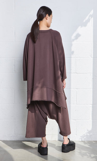 Plümo- In sunlight, person faces a white wall in a relaxed brown Kelie Sweater and pants with black platform shoes.
