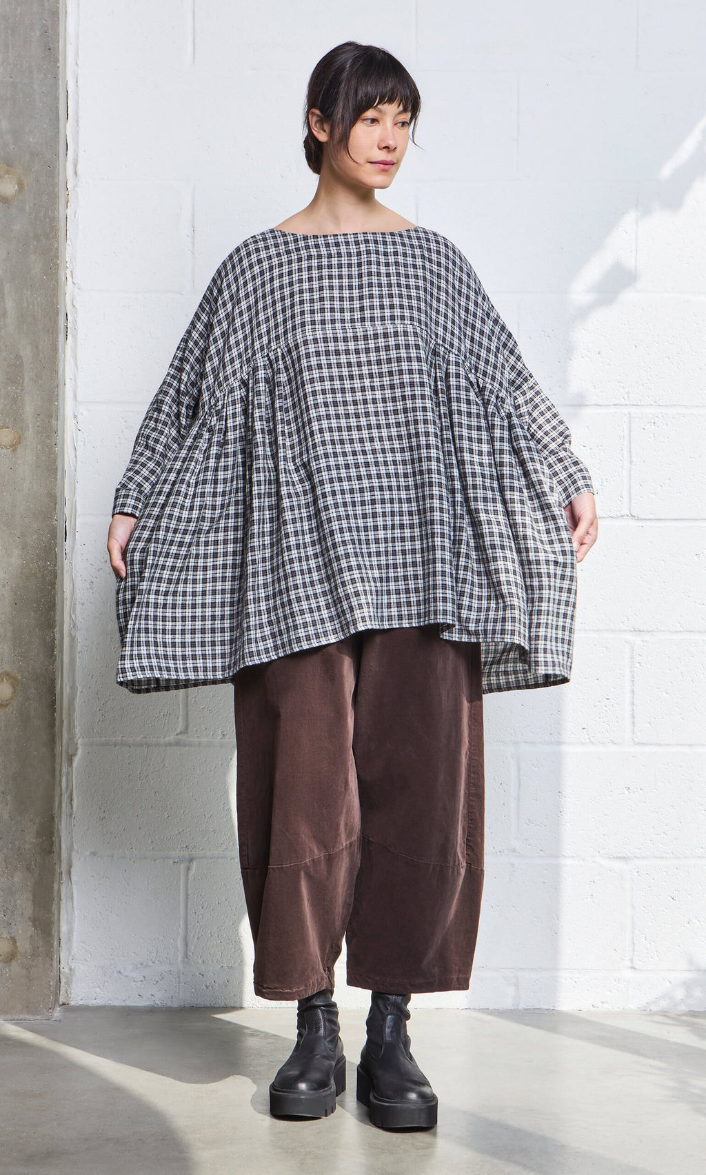 Plümo- Person wears the Kelli Blouse - Plaid with wide brown pants and black platform shoes, standing indoors.