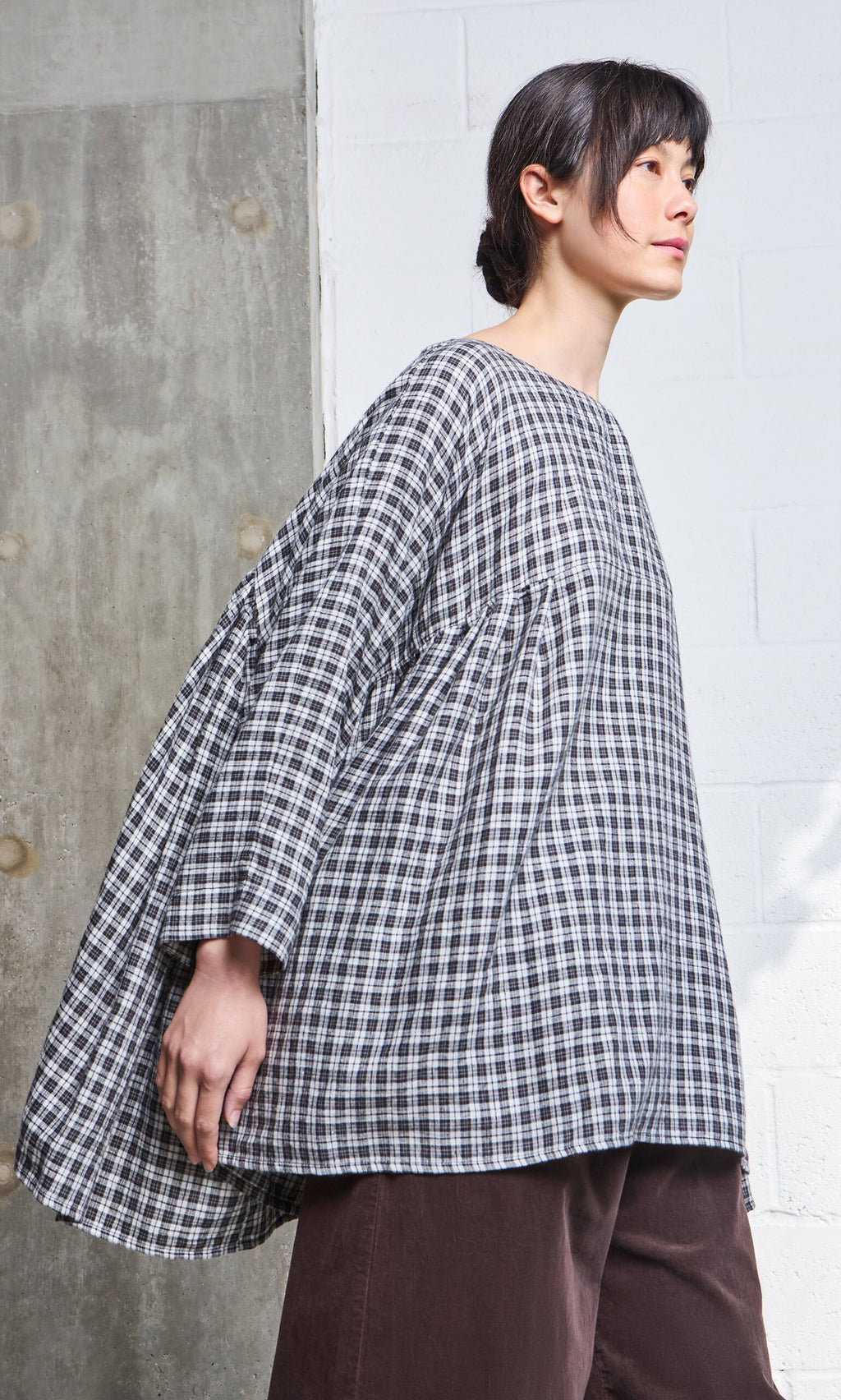 Plümo- A person in the Kelli Blouse - Plaid and brown pants stands against a textured wall.