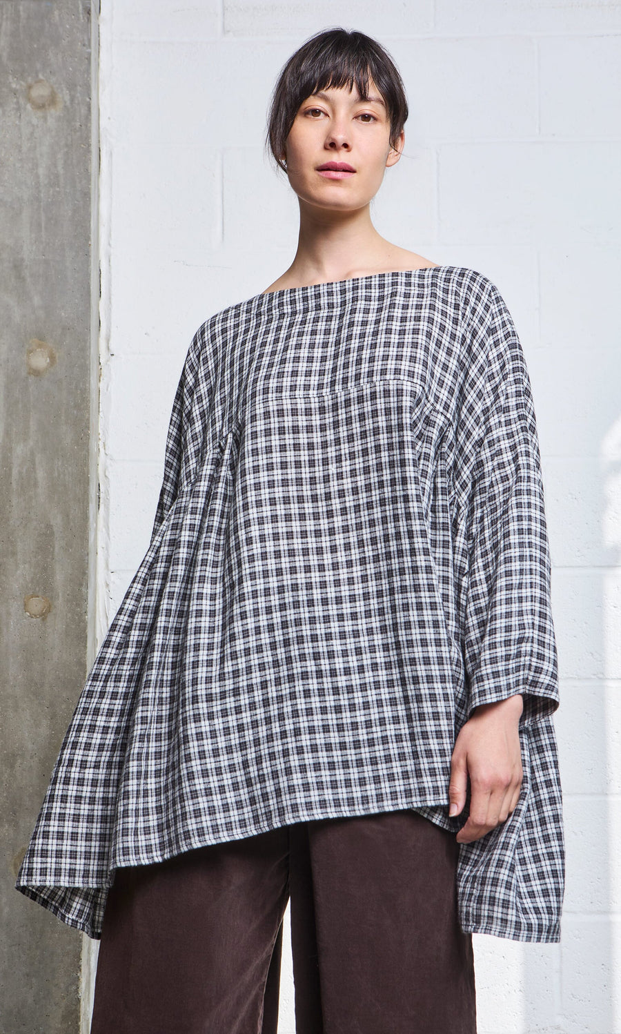 Plümo- Woman wearing the Kelli Blouse - Plaid with wide sleeves and dark brown pants, standing indoors.