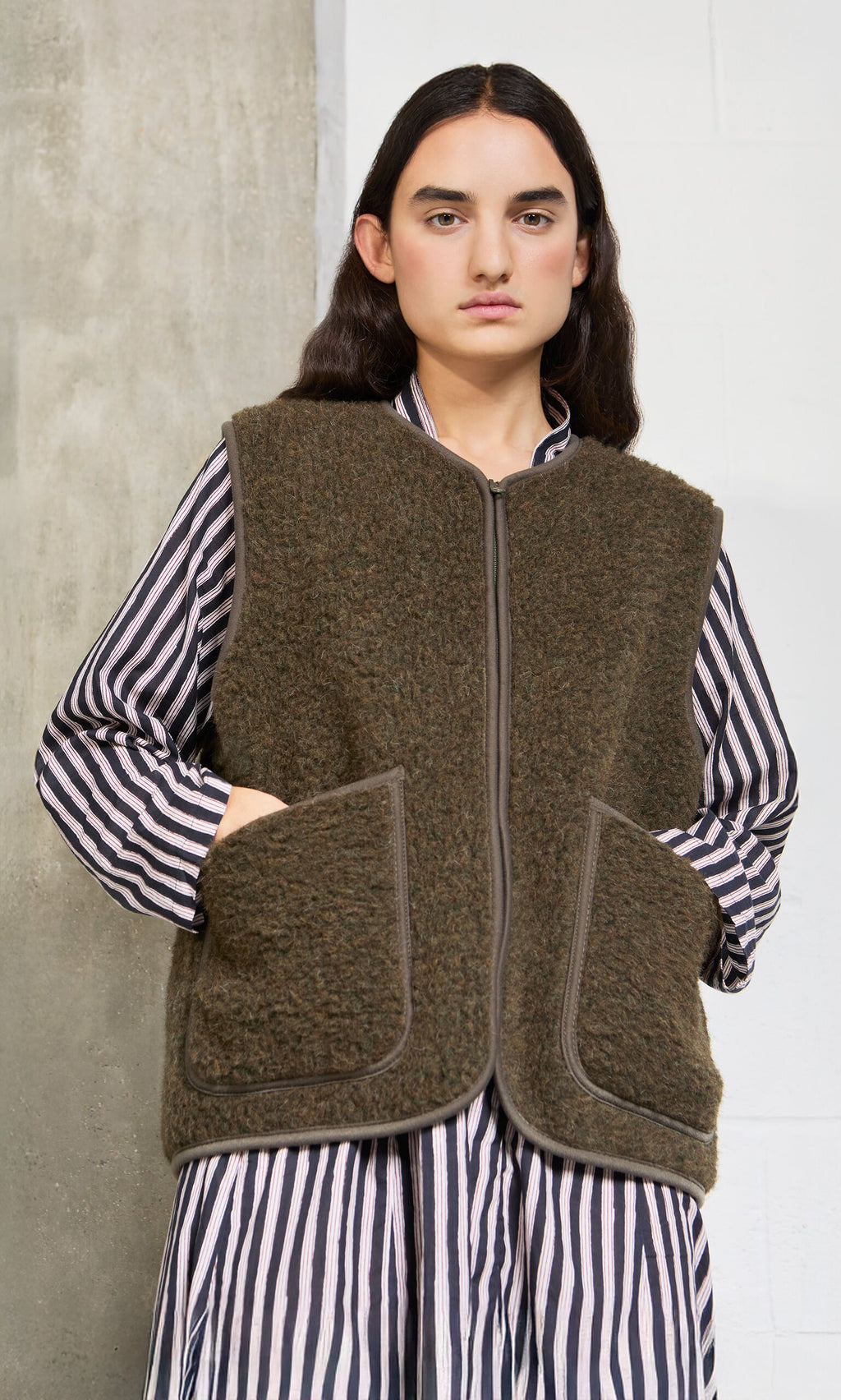 Plümo- Woman in a striped dress wears the Khaki Wool Gilet with large pockets and cropped fit, standing by a concrete wall.