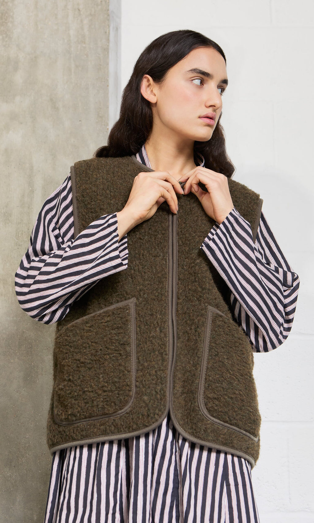 Plümo- Woman in a striped shirt and cropped Khaki Wool Gilet stands against a concrete and white wall, looking to the side.