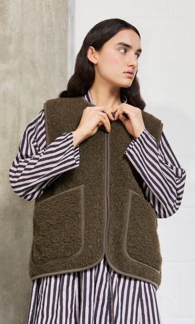 Plümo- Woman in a striped shirt and cropped Khaki Wool Gilet stands against a concrete and white wall, looking to the side.