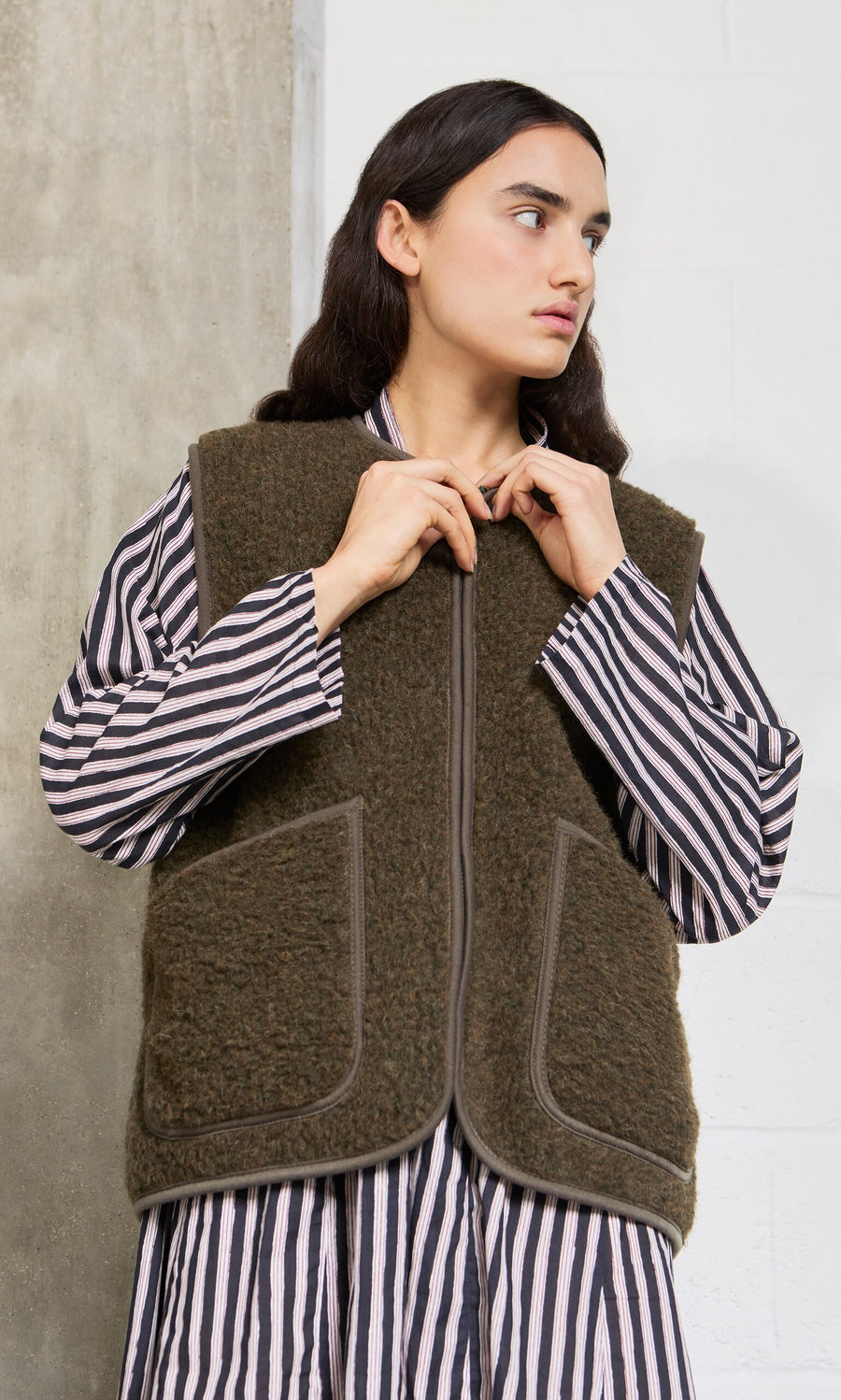 Plümo- Woman in a striped shirt and cropped Khaki Wool Gilet stands against a concrete and white wall, looking to the side.