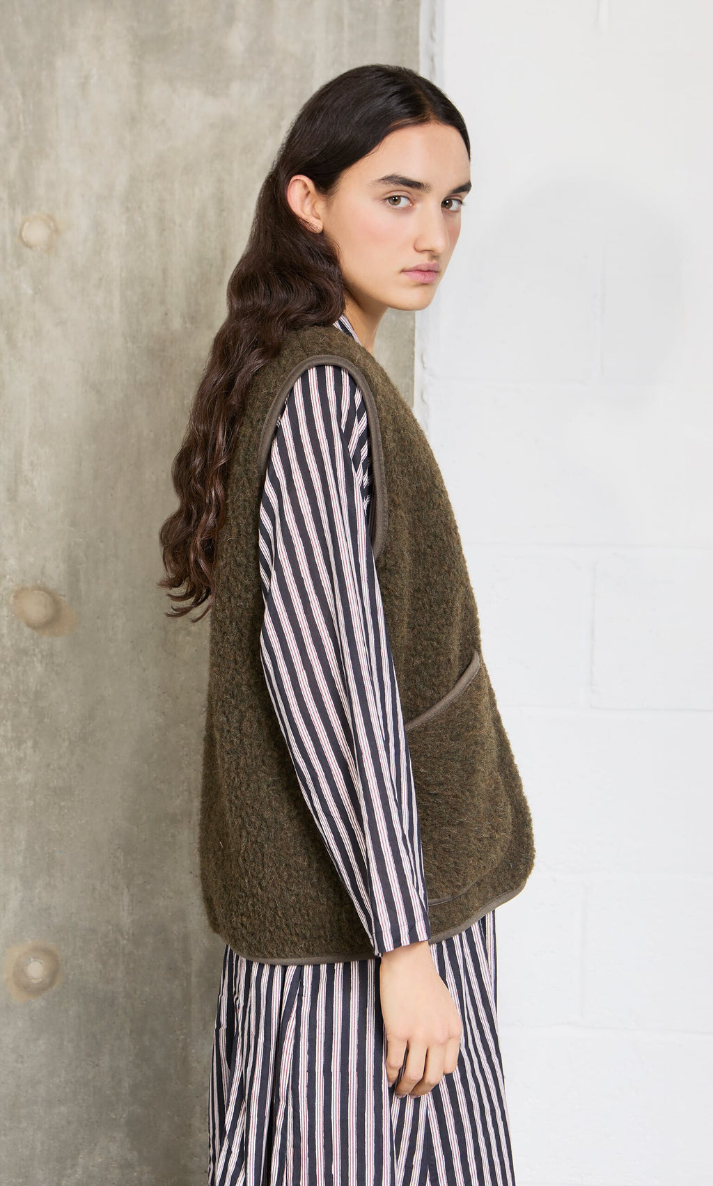 Plümo- Woman in a striped dress and Khaki Wool Gilet stands sideways against a concrete and white wall.