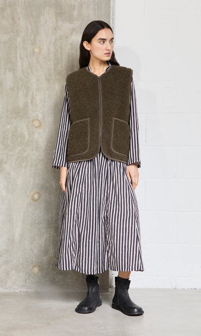 Plümo- A person in a striped dress, khaki wool gilet, and black boots stands against a concrete and white wall.