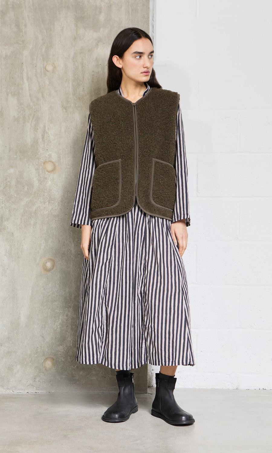 Plümo- A person in a striped dress, khaki wool gilet, and black boots stands against a concrete and white wall.