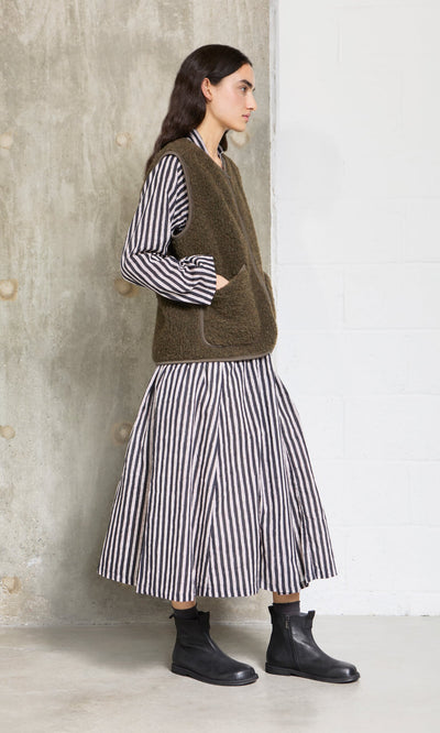 Plümo- Woman in the Hoyland Dress - Charcoal Stripe, brown vest, and black ankle boots stands by a textured wall.