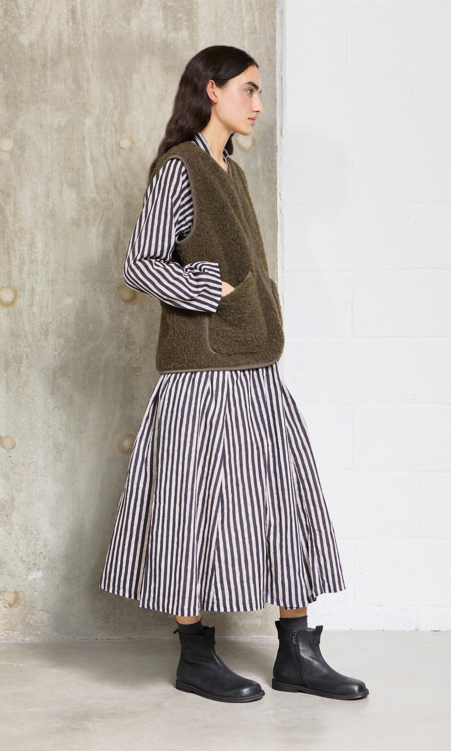 Plümo- Woman in a striped dress, Khaki Wool Gilet, and black ankle boots stands by a concrete and white wall.