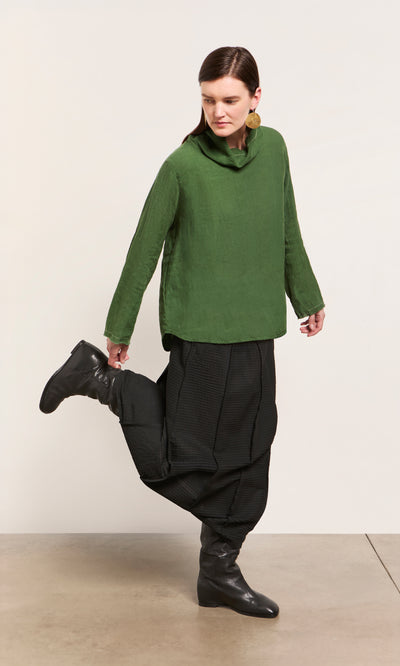 Plümo  Woman in Kim Ruffle Stitch Blouse, black pants, and boots poses with one leg bent on a light floor against a white wall.