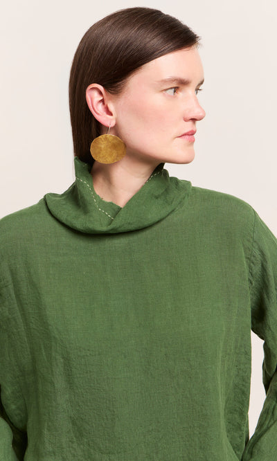 Plümo  Woman in a Kim Ruffle Stitch Blouse, green linen with cowl neck, large gold earring, gazing thoughtfully sideways.