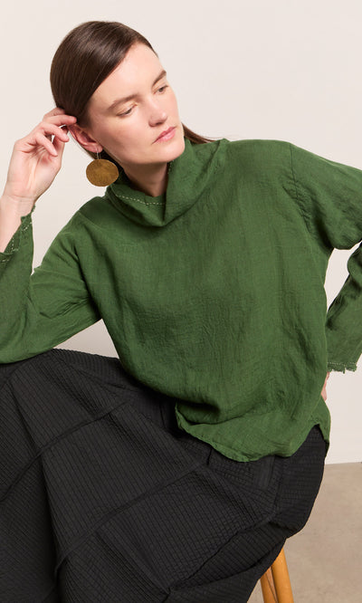 Plümo  Woman wearing the Kim Ruffle Stitch Blouse in green, seated with a large gold earring against a neutral background.