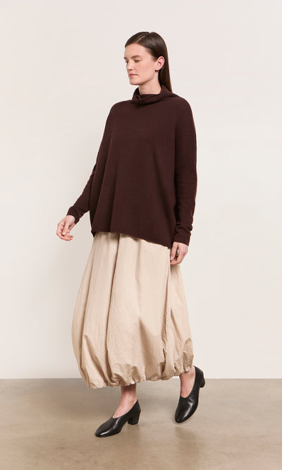 Plümo  Woman in the Kloster Rollneck merino yak sweater, beige balloon skirt, black shoes, plain background.