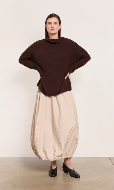 Plümo  A woman wears the Kloster Rollneck in brown ochre and a beige skirt with black heels, standing by a white wall.