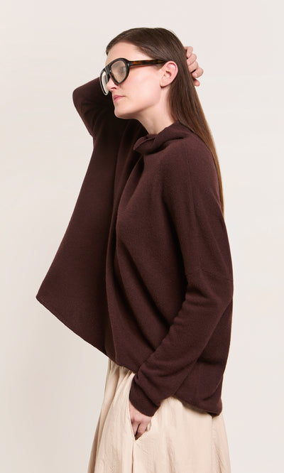 Plümo  Person in Kloster Rollneck, beige skirt, and black glasses stands sideways, hand on head, against plain background.