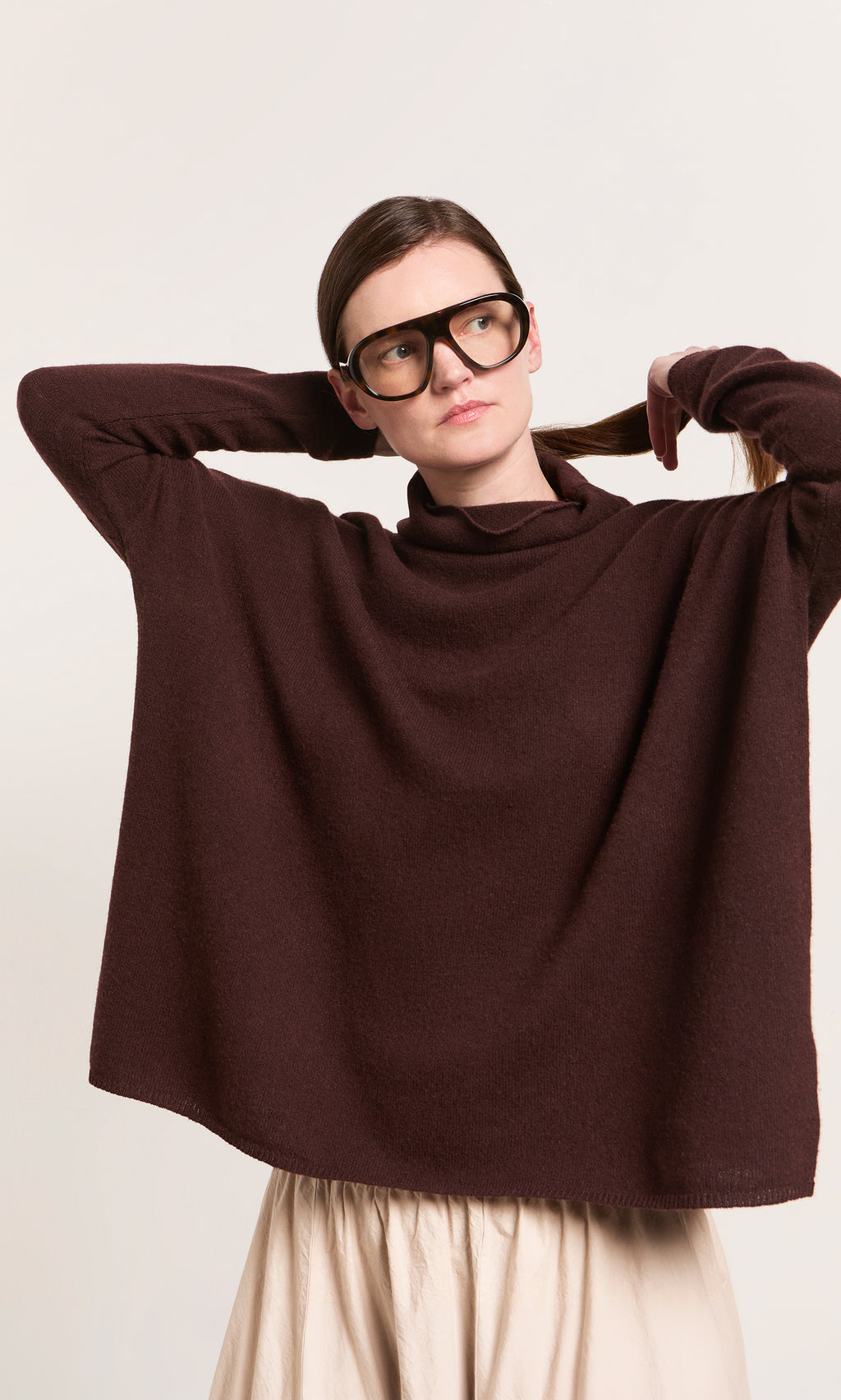 Plümo  Woman in black glasses and cream skirt wears Kloster Rollneck, arms raised, holding her hair back.
