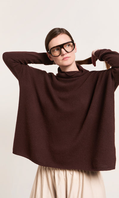 Plümo  Woman in black glasses and cream skirt wears Kloster Rollneck, arms raised, holding her hair back.