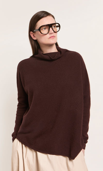 Plümo  Woman wears oversized black glasses, a Kloster Rollneck sweater in brown ochre, and a beige skirt against a plain background.