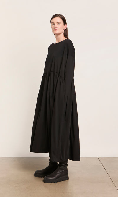 Plümo  Woman in the Kobke Dress—an organic cotton jersey black maxi—pairs it with chunky boots against a plain wall.