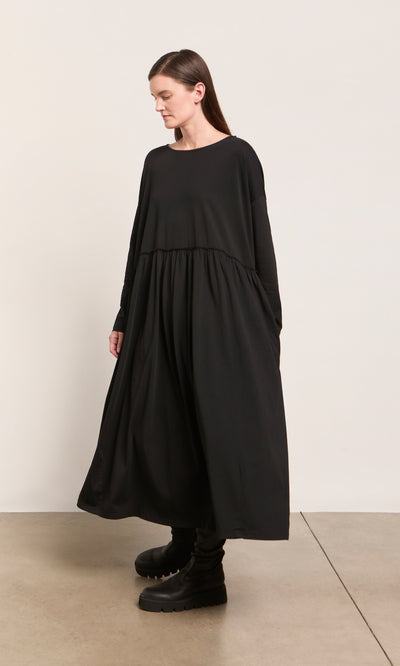 Plümo  Woman in the Kobke Dress—loose, black, organic cotton jersey—paired with chunky boots by a cream wall.