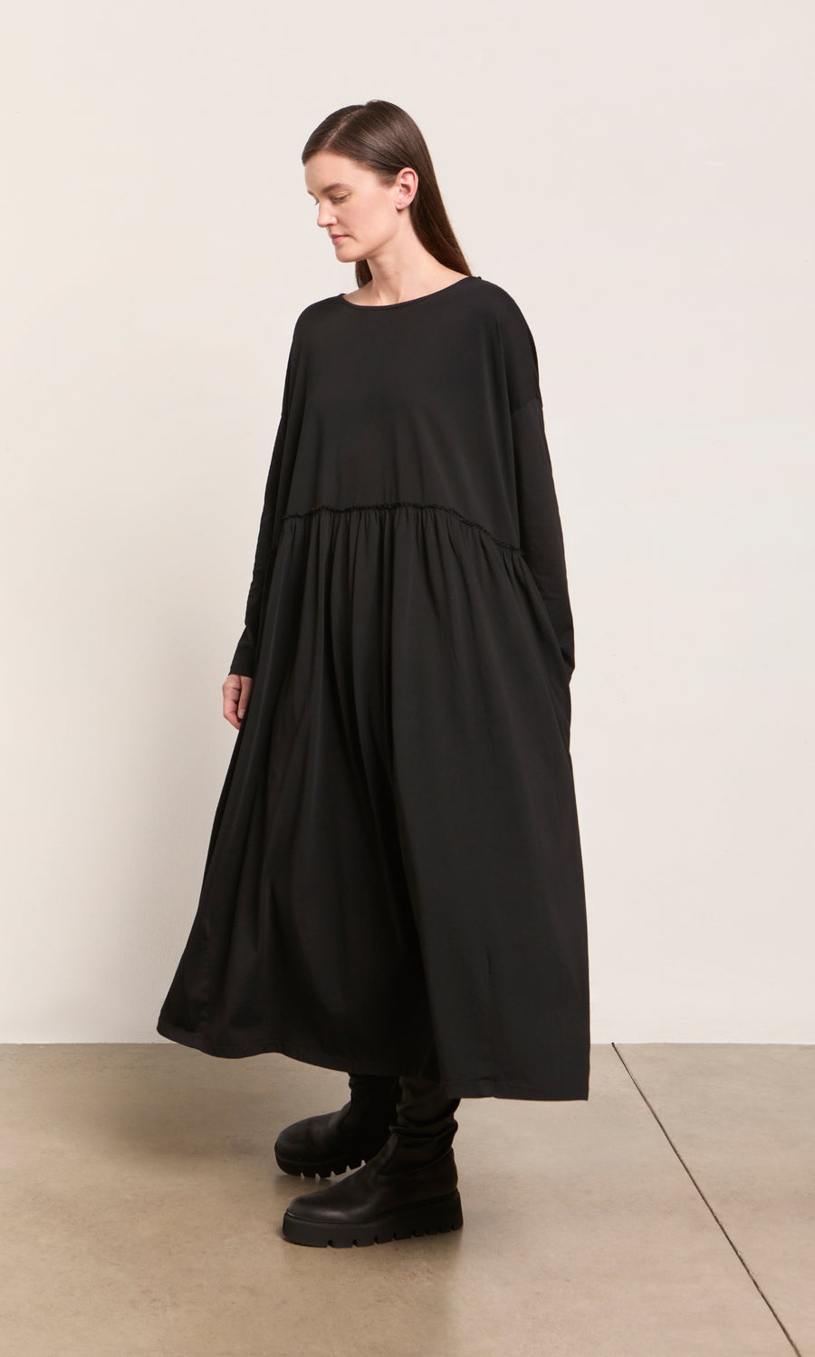Plümo  Woman in the Kobke Dress—loose, black, organic cotton jersey—paired with chunky boots by a cream wall.