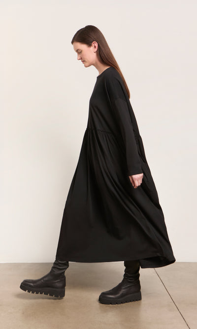 Plümo  Woman wears the Kobke Dress—loose black organic cotton jersey—paired with chunky boots, walking indoors.