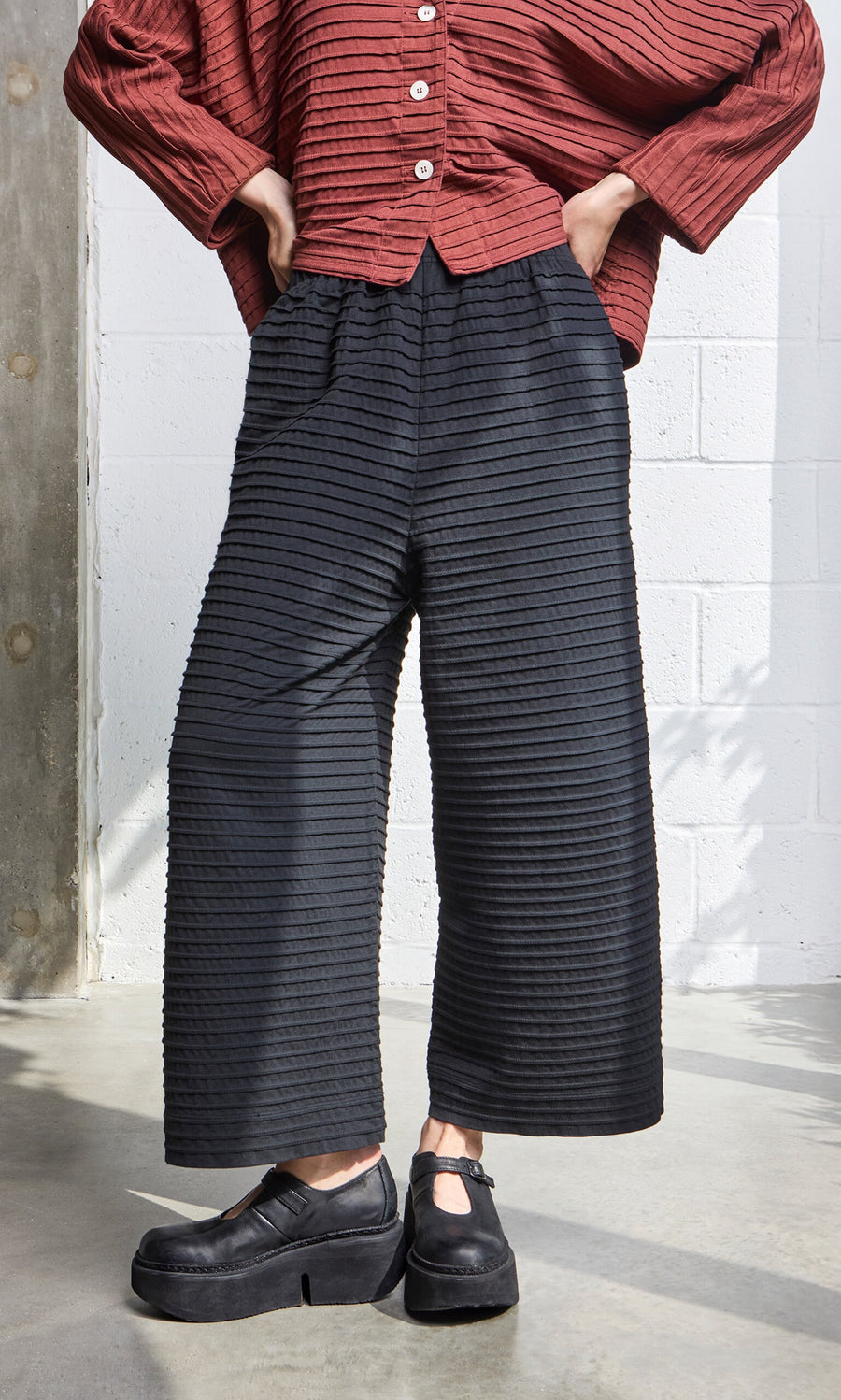 Plümo- Person wearing a red textured top, Koji Pants in black Japanese design, and black platform shoes, standing indoors.