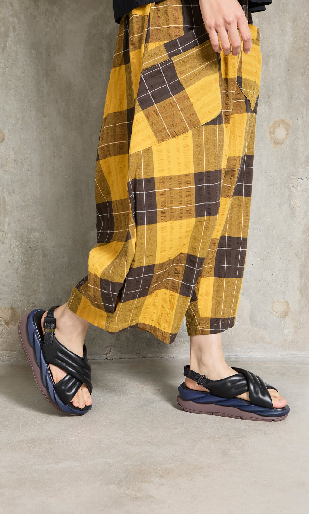 Plümo- Person in yellow and brown plaid pants walks on gray concrete floor wearing Korra Sandals.