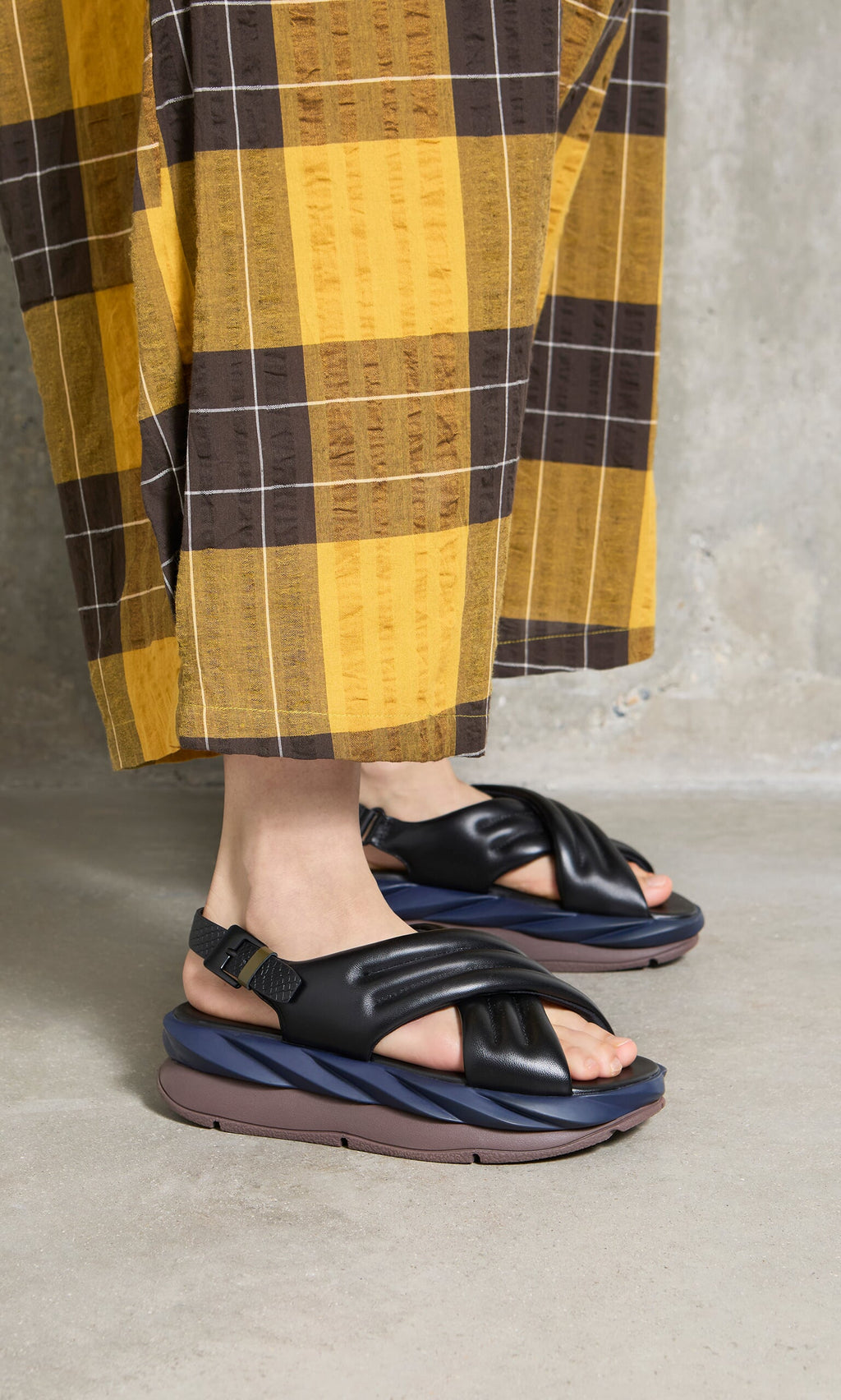 Plümo- Person wearing yellow and brown plaid pants with black Korra Sandals featuring adjustable buckle strap on concrete floor.