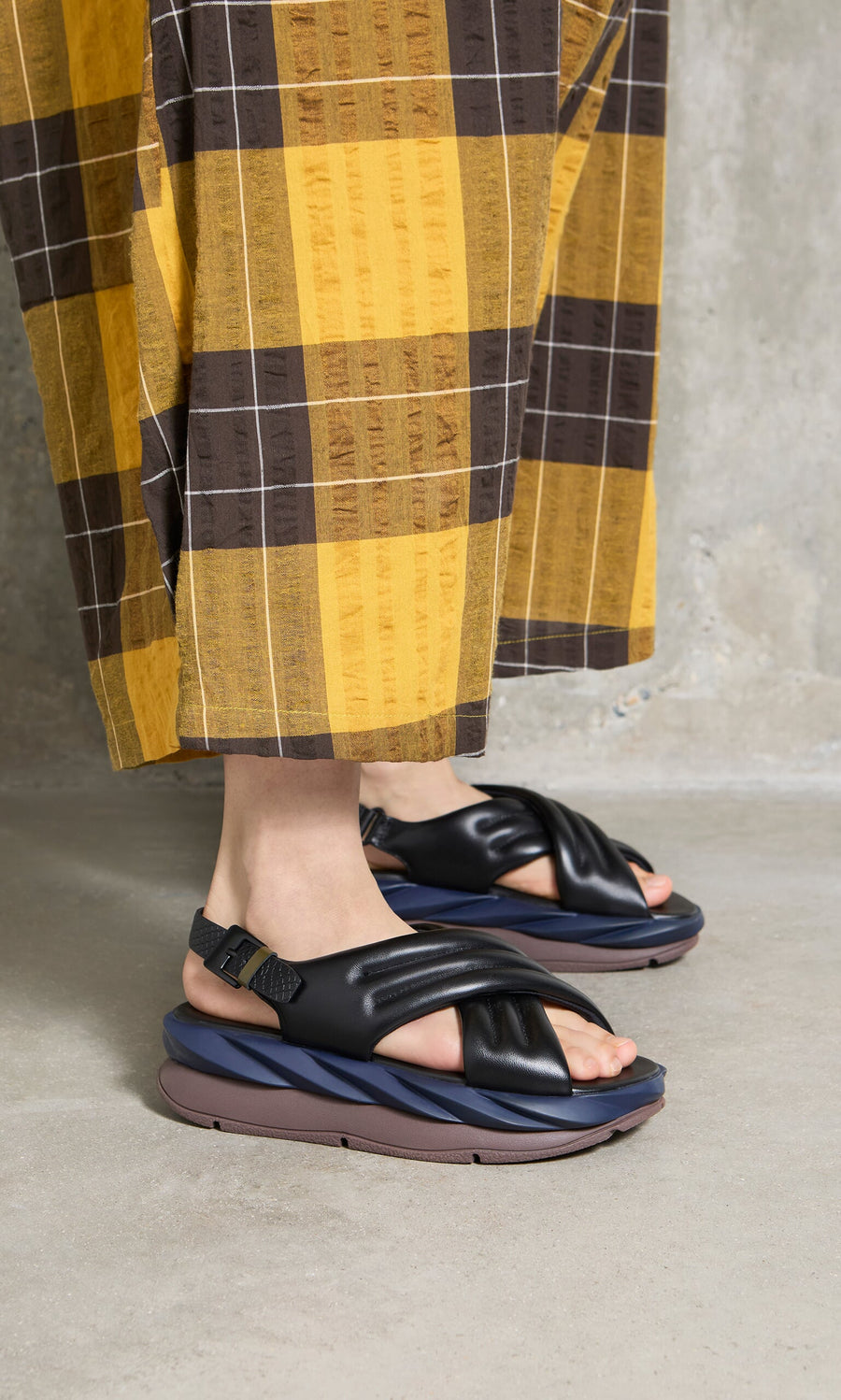 Plümo- Person wearing yellow and brown plaid pants with black Korra Sandals featuring adjustable buckle strap on concrete floor.