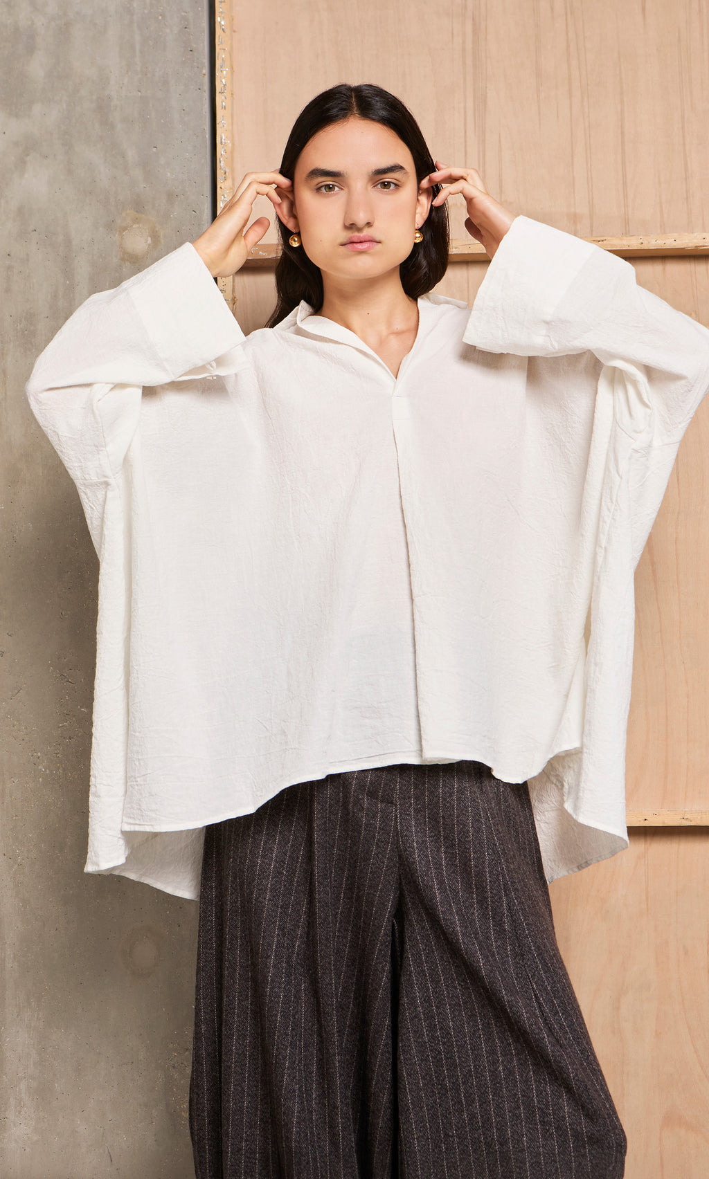 Plümo  Person in a loose Laundered Shirt stands against a wooden and concrete background.