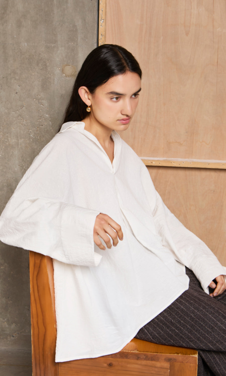 Plümo  Woman in Laundered Shirt sits on wooden chair, neutral background, gazing thoughtfully to the side.