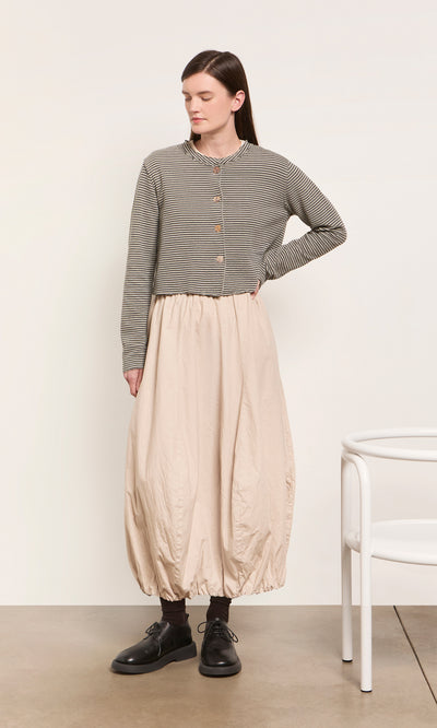 Plümo  Woman in the Lavelle Top and loose beige skirt stands by a white chair on a plain background.