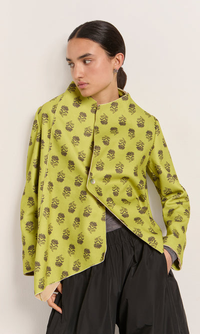 Plümo  Woman in Leandra Jacket and black pants stands by a white wall, looking left.