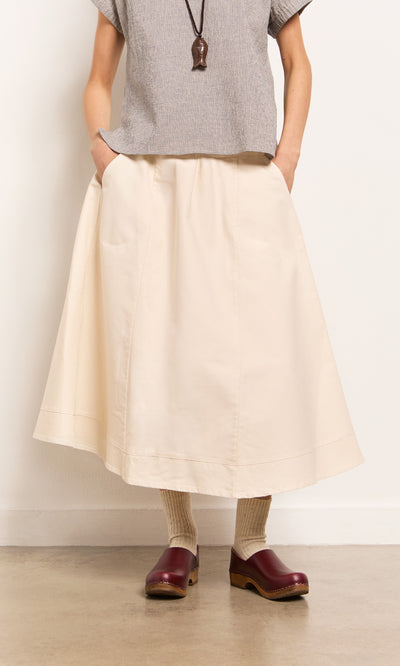Plümo- A person in the Lee Skirt, gray top, beige socks, and burgundy clogs stands against a plain wall.