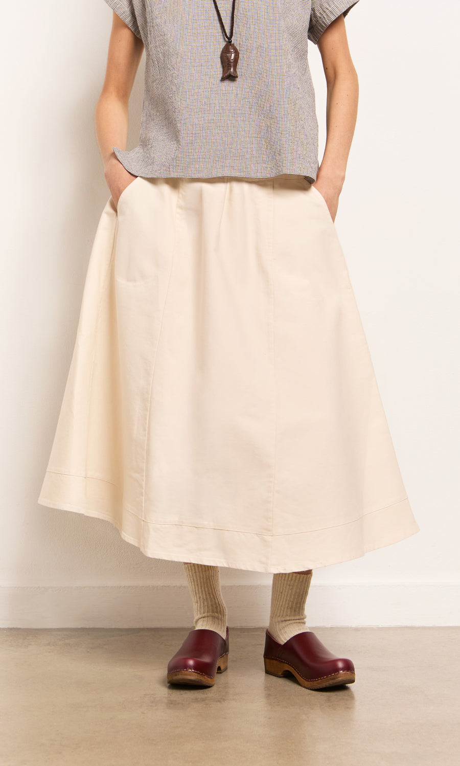 Plümo- A person in the Lee Skirt, gray top, beige socks, and burgundy clogs stands against a plain wall.