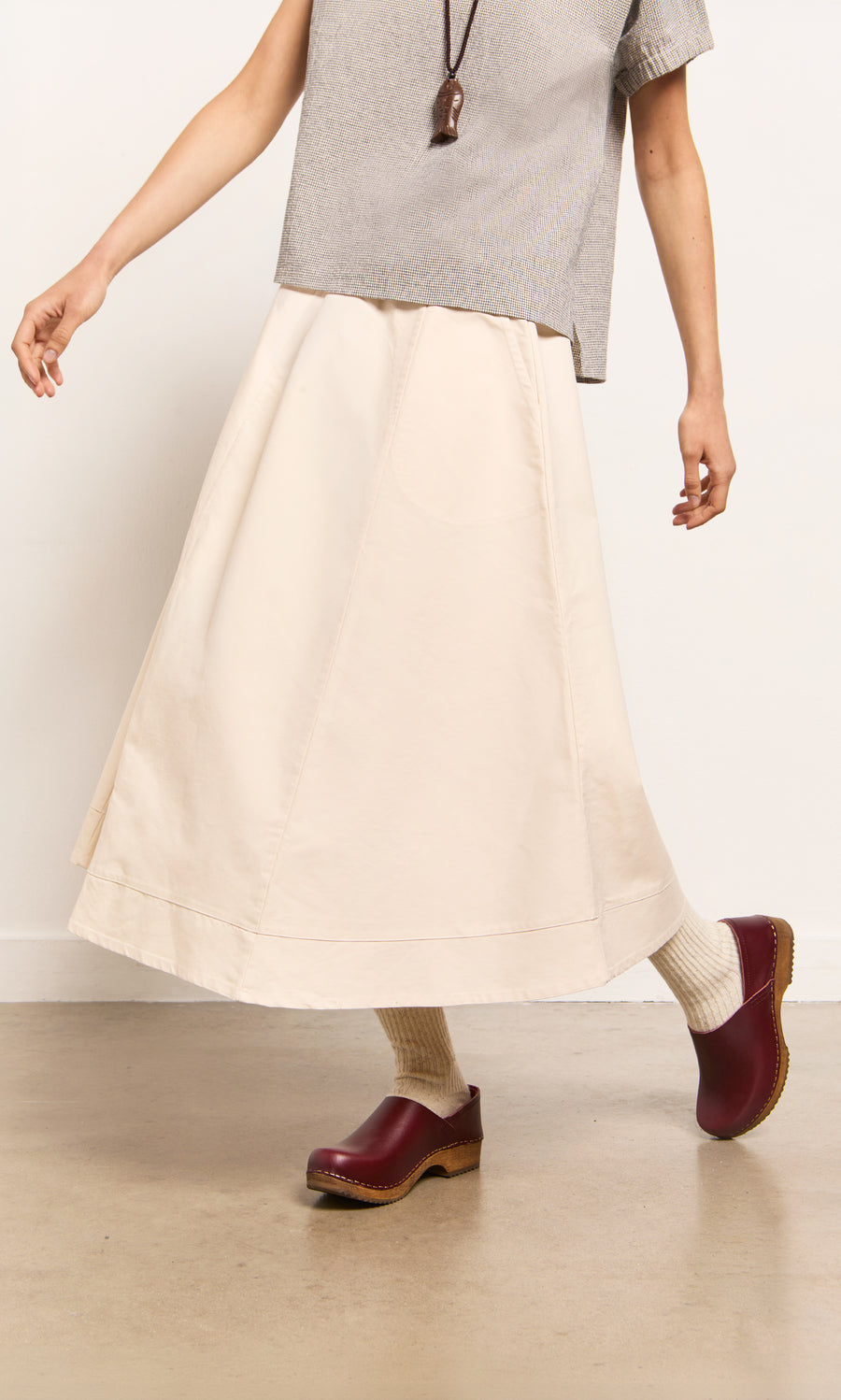 Plümo- Model in a beige Lee Skirt, gray top, cream socks, and burgundy clogs walks indoors against a plain background.