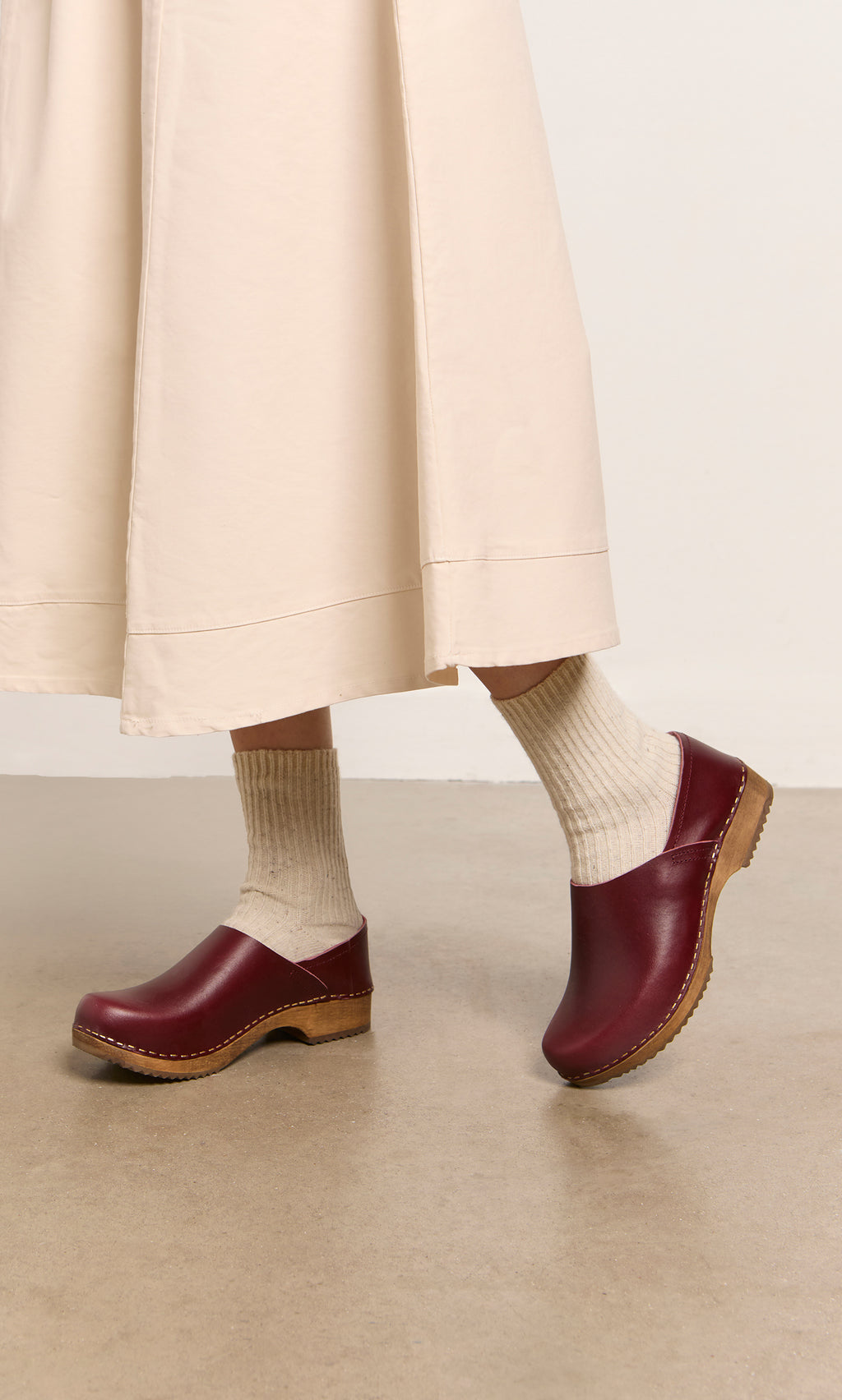 Plümo  Wearing cream socks and Low Jord Clogs, a person stands on a tan floor in a light beige midi-length skirt.